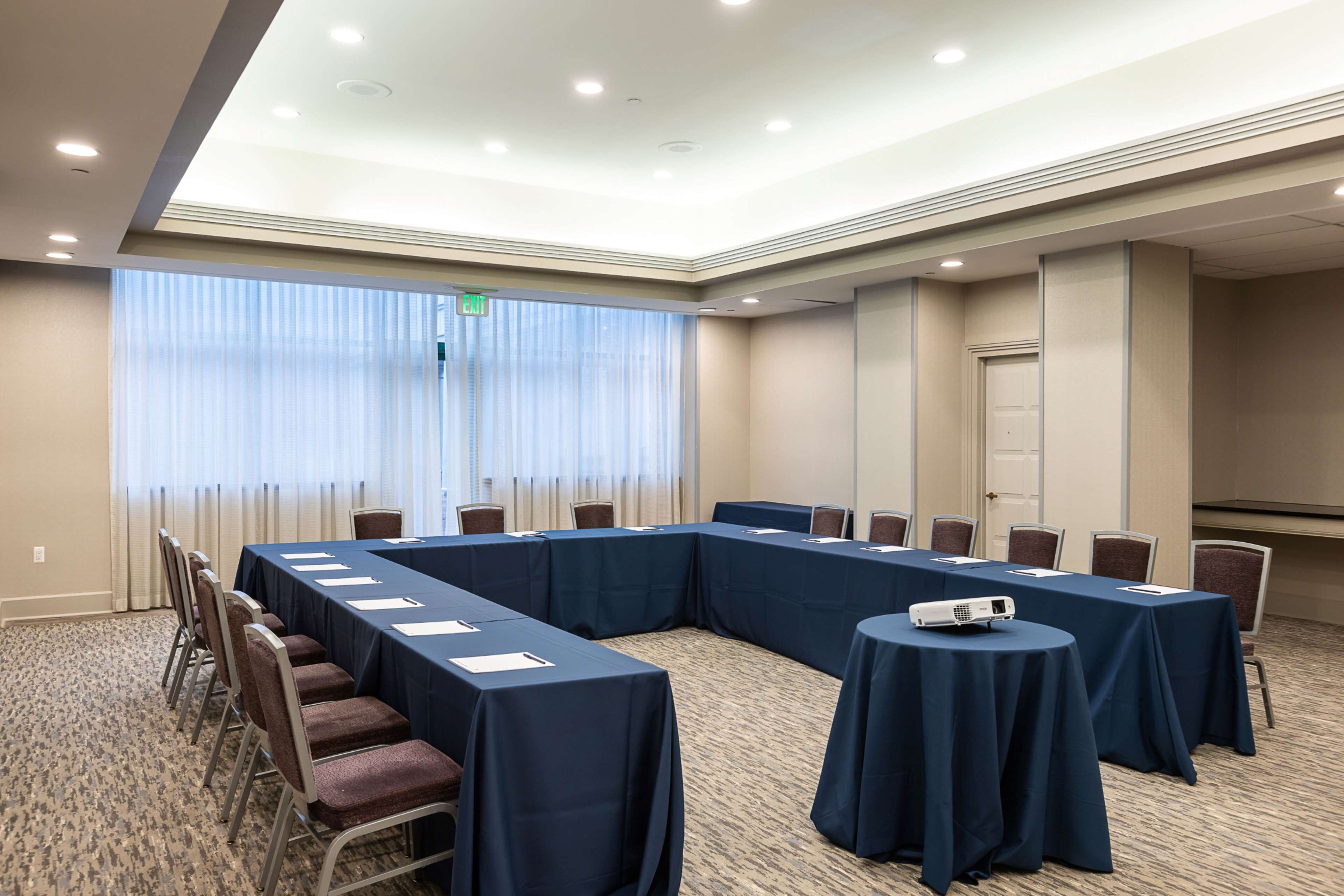 Un salón de conferencias con una larga mesa, sillas, un proyector y un amplio ventanal con cortinas.