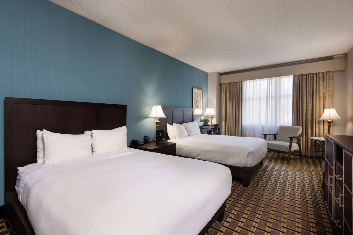A hotel room with two beds, a desk, a chair, and a large window with curtains.