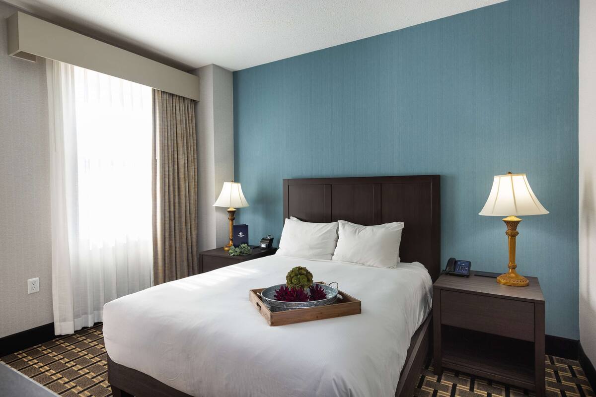 A hotel room with a large bed, two bedside tables with lamps, a tray with flowers, and a view of the outside through a window with curtains.