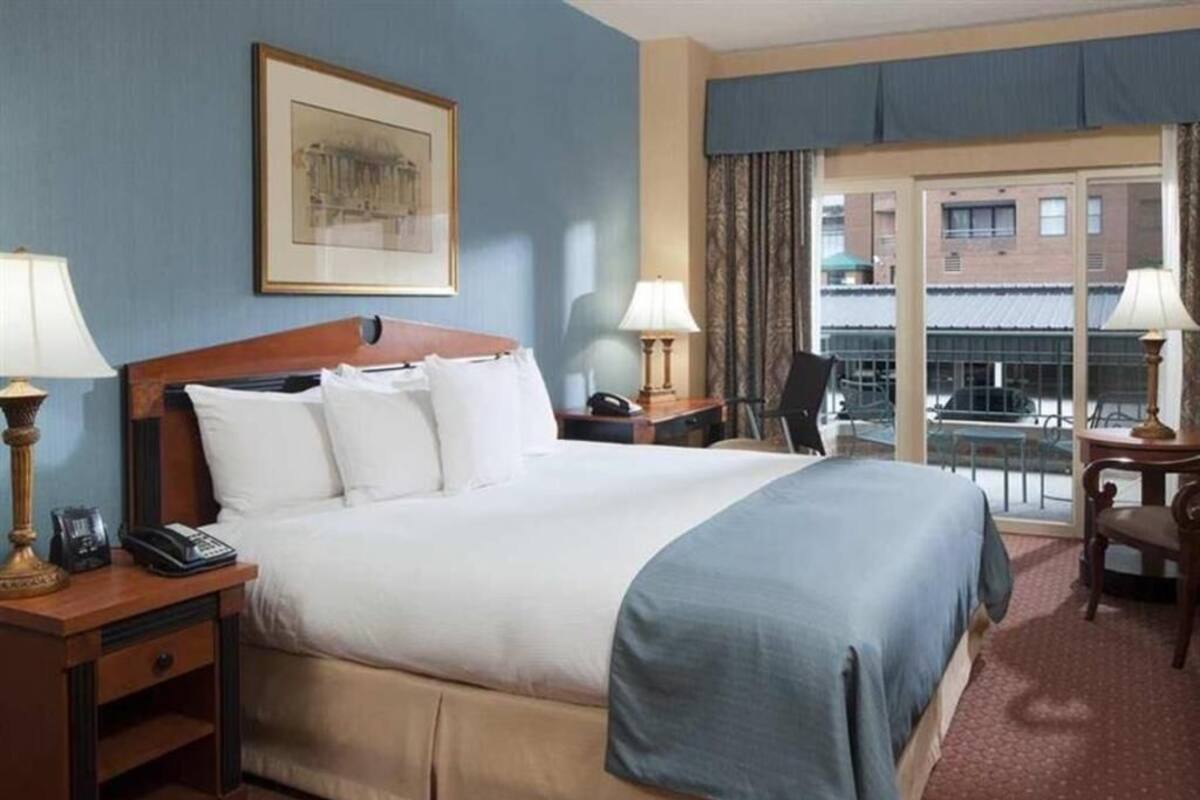 A hotel room with a large bed, two bedside tables with lamps, a chair, a desk, and a window with a view of buildings.