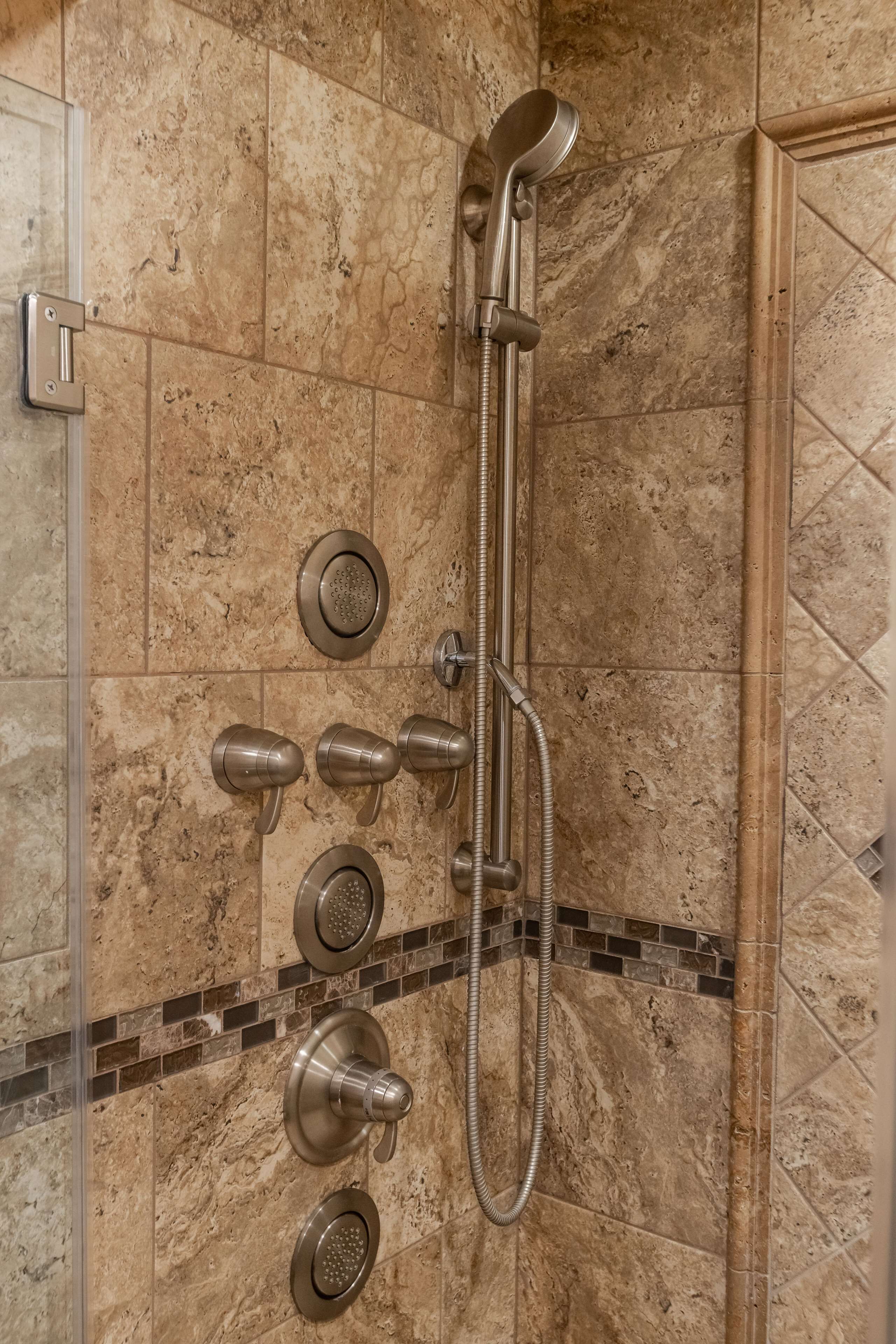 Ducha con varios cabezales y un cabezal de ducha manual, frente a una pared de azulejos con un borde de mosaico.