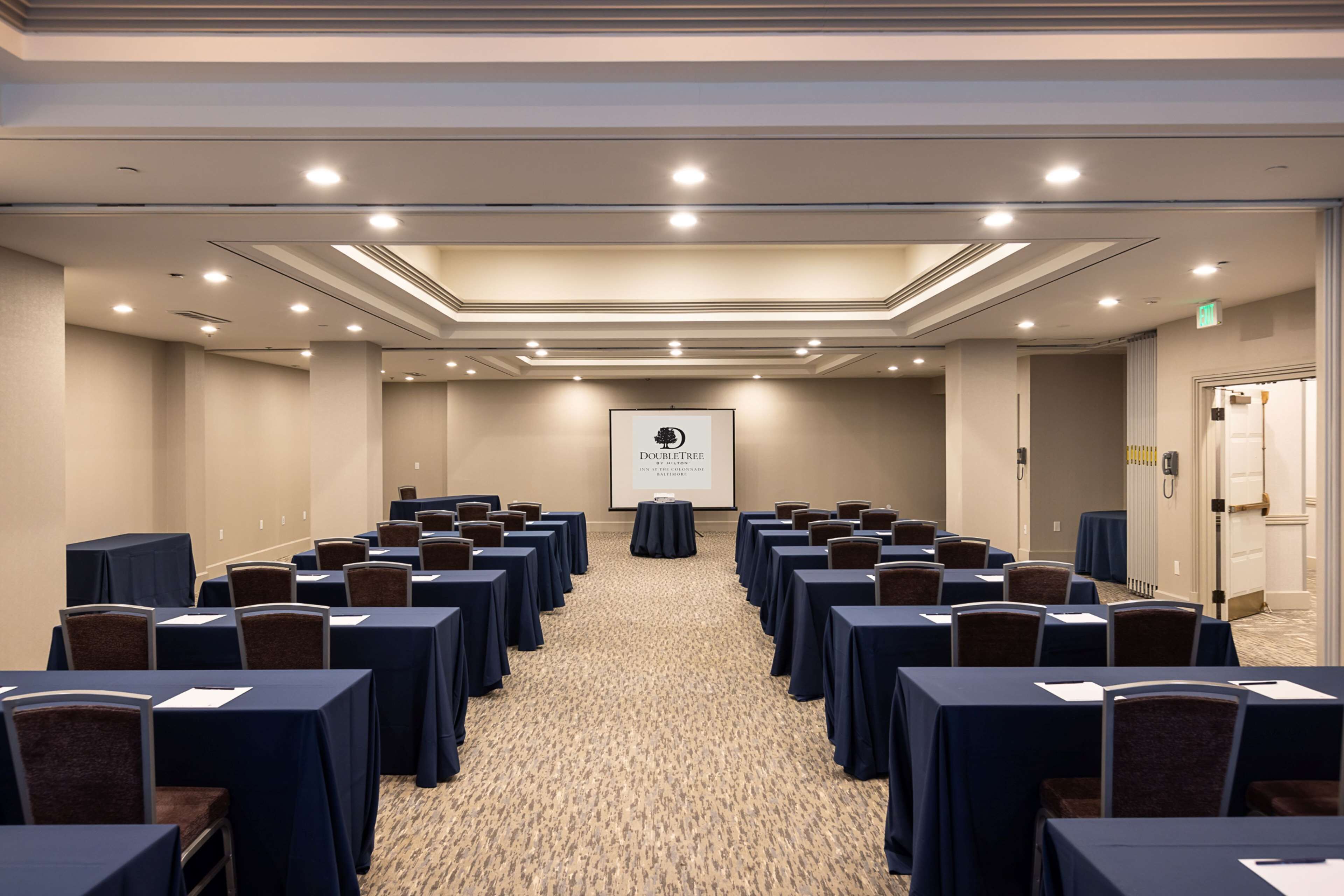 Un salón de conferencias con filas de mesas y sillas, piso alfombrado y una pantalla proyectora mostrando "DOUBLETREE".