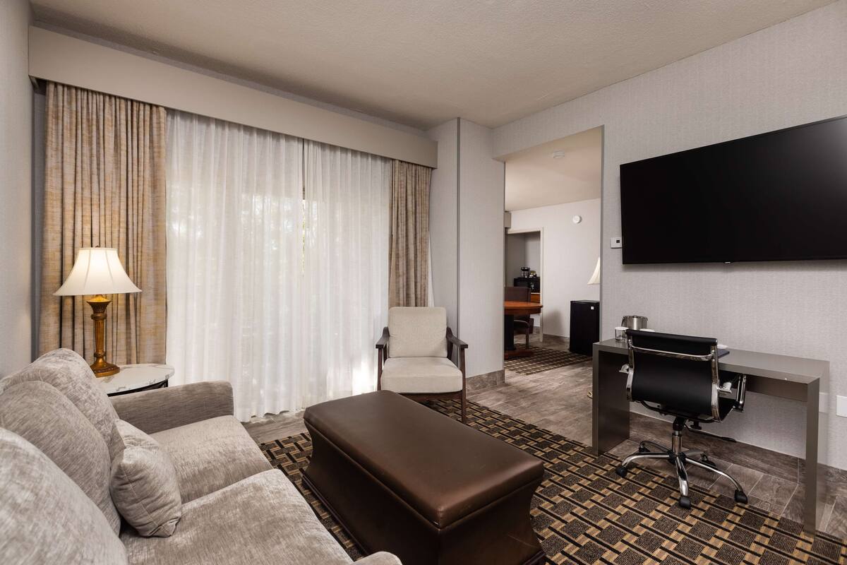 A hotel room with a sofa, a brown ottoman, a desk with a chair, a flat-screen TV, and a large window with curtains.