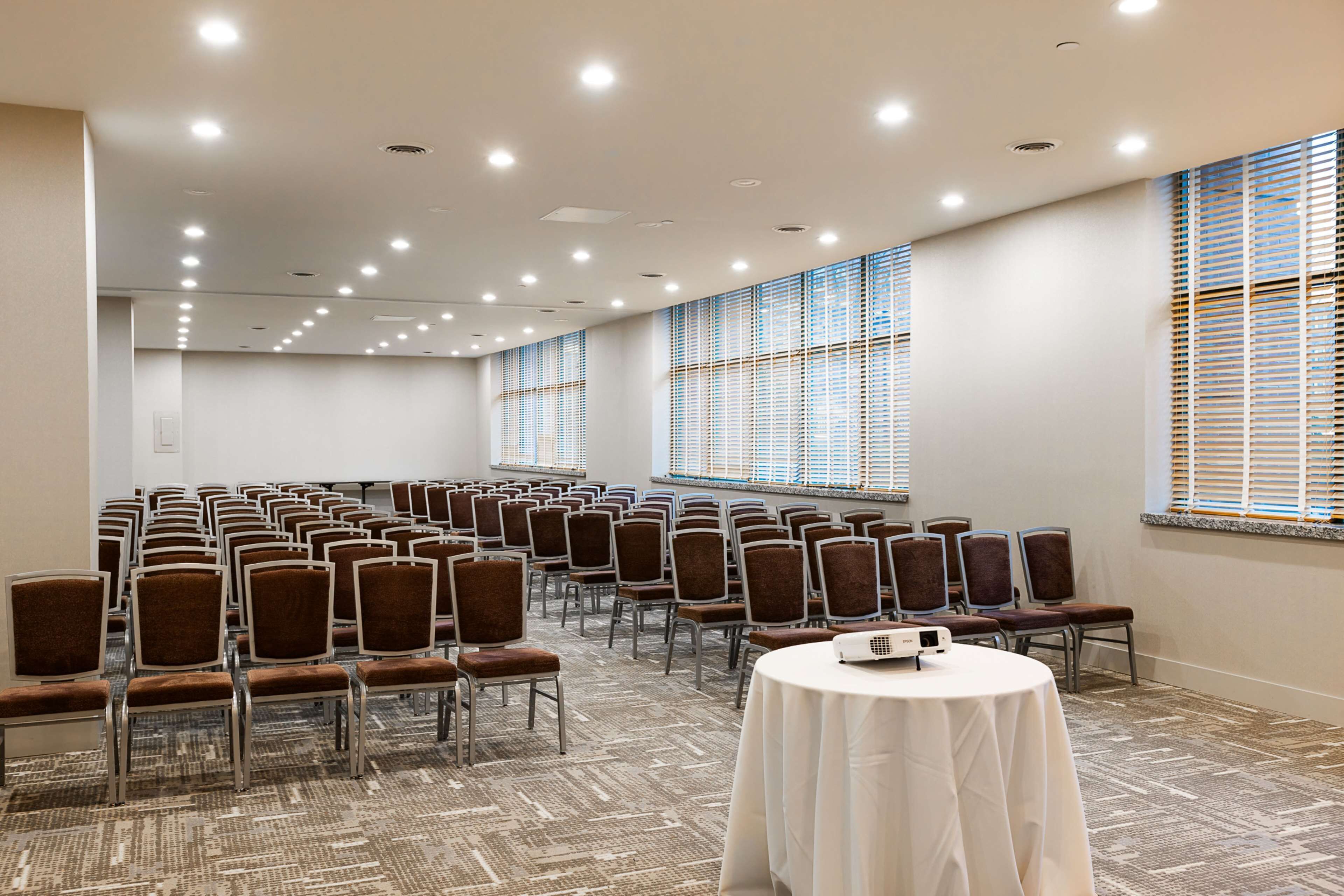 Une salle de conférence avec des rangées de chaises, un projecteur posé sur une table et de grandes fenêtres équipées de stores.
