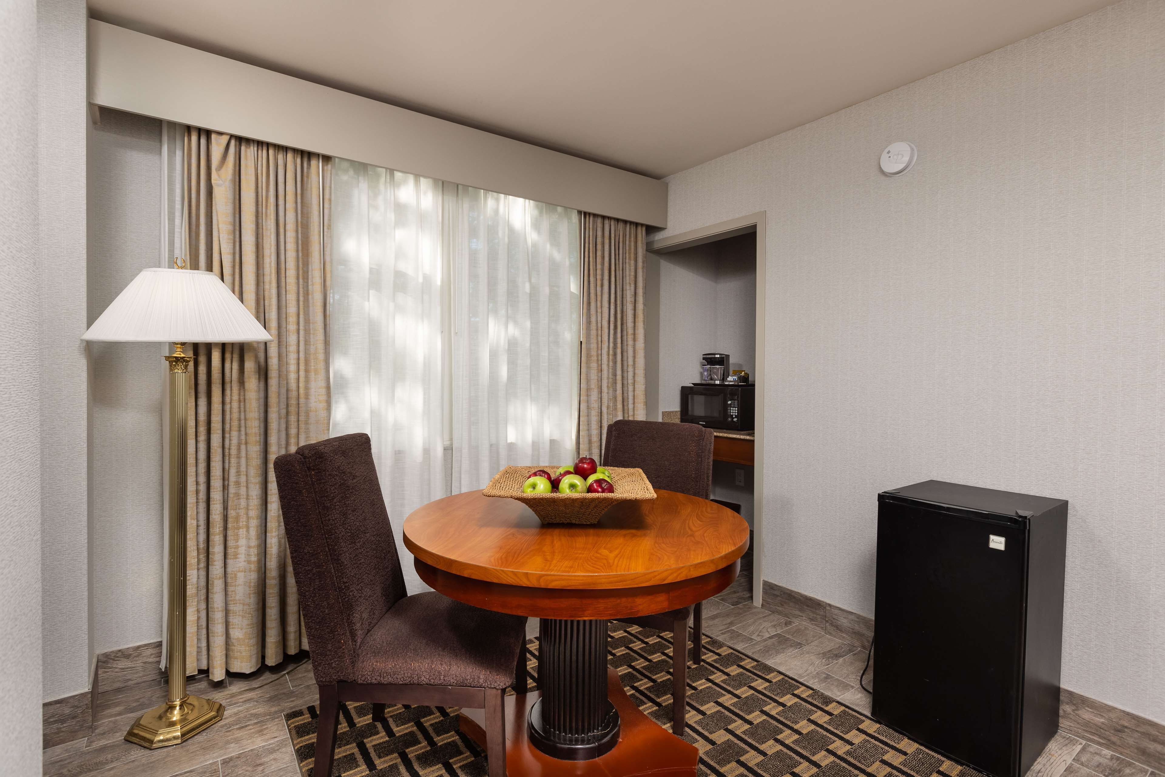 A hotel room with a round wooden dining table, two chairs, a small black refrigerator, a lamp, and a fruit basket.