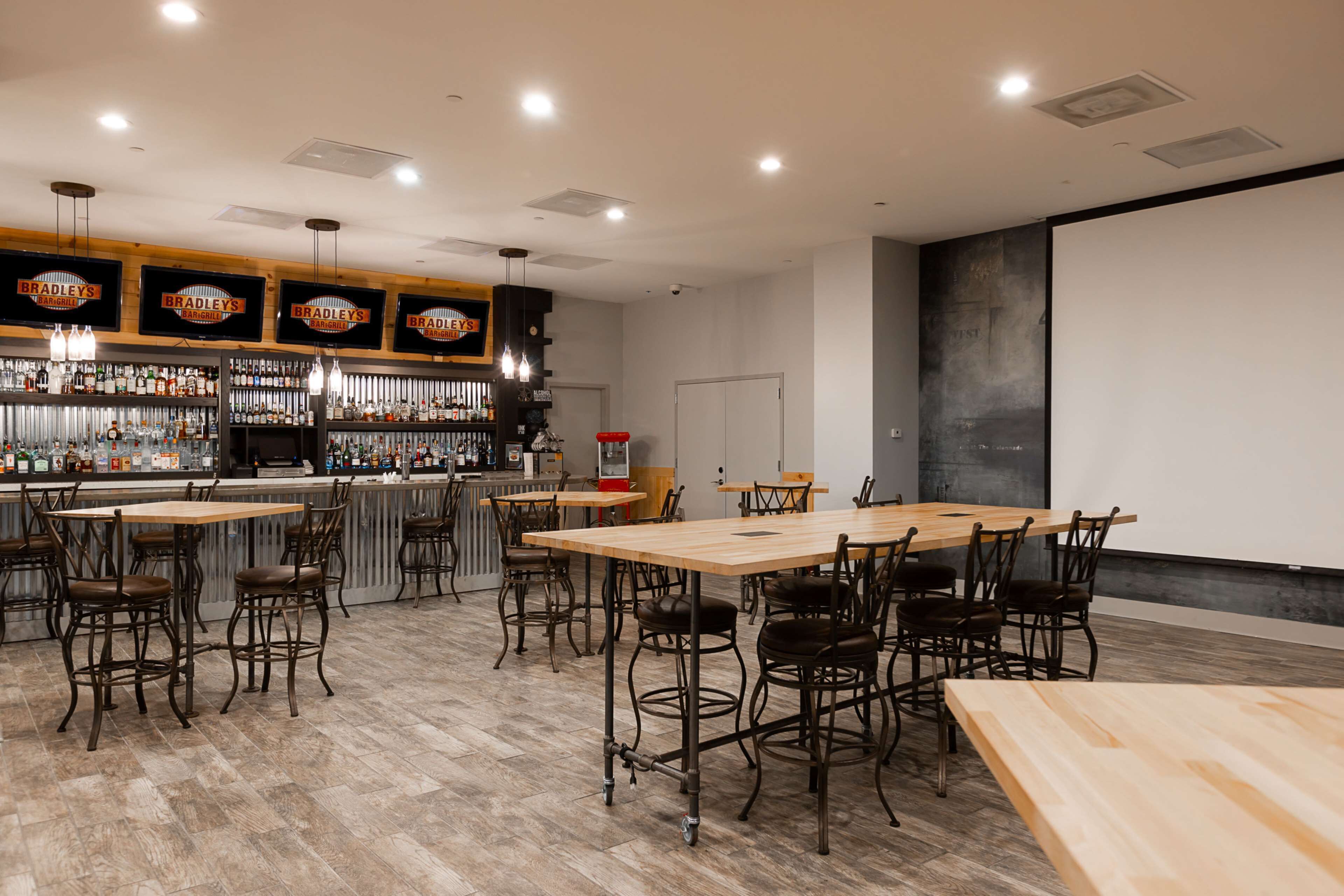 A bar area with multiple tables and chairs, a bar counter with various bottles, and a projector screen on the wall.