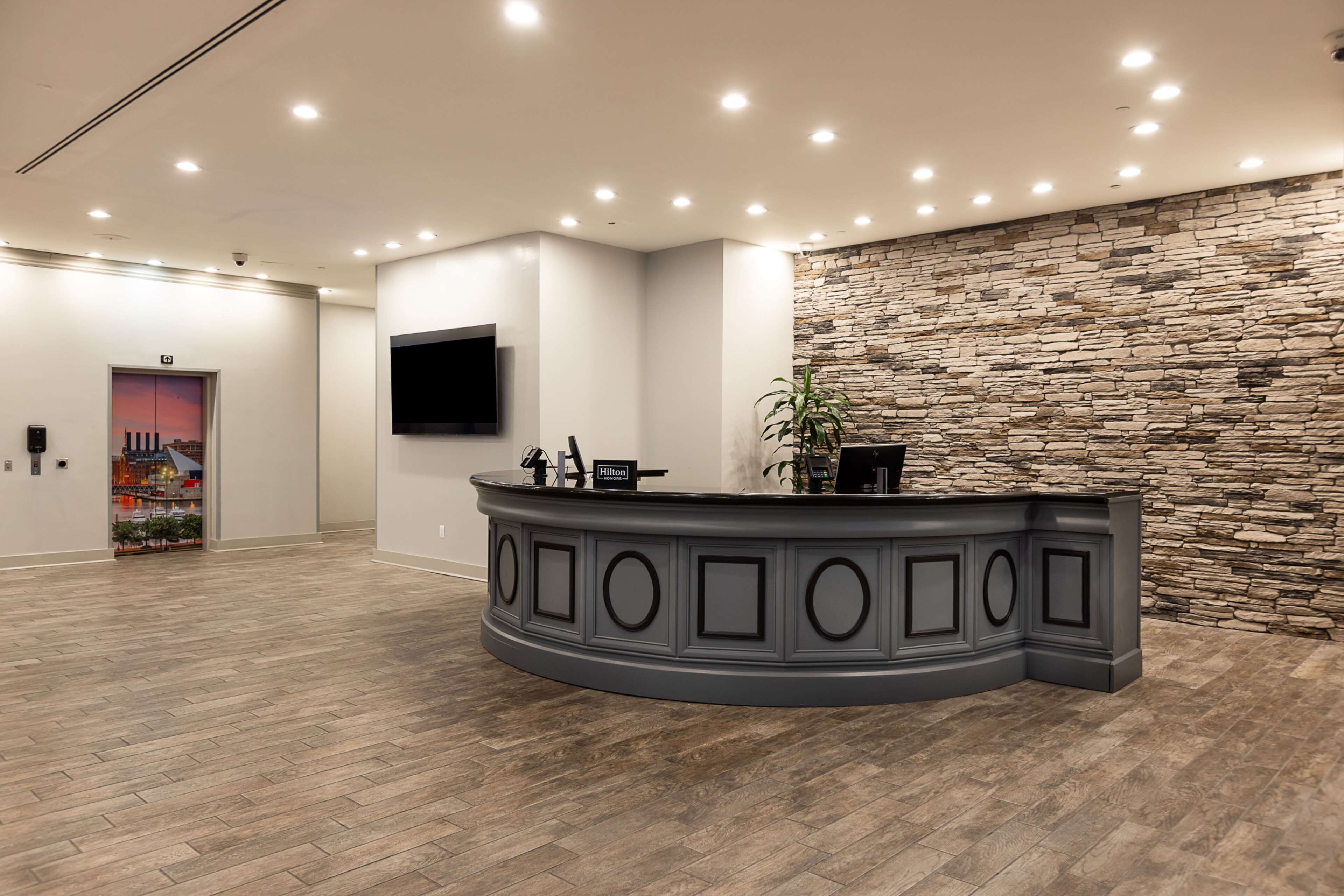 A hotel reception area with a curved grey reception desk, a flat-screen TV mounted on the wall, and a stone wall accent.