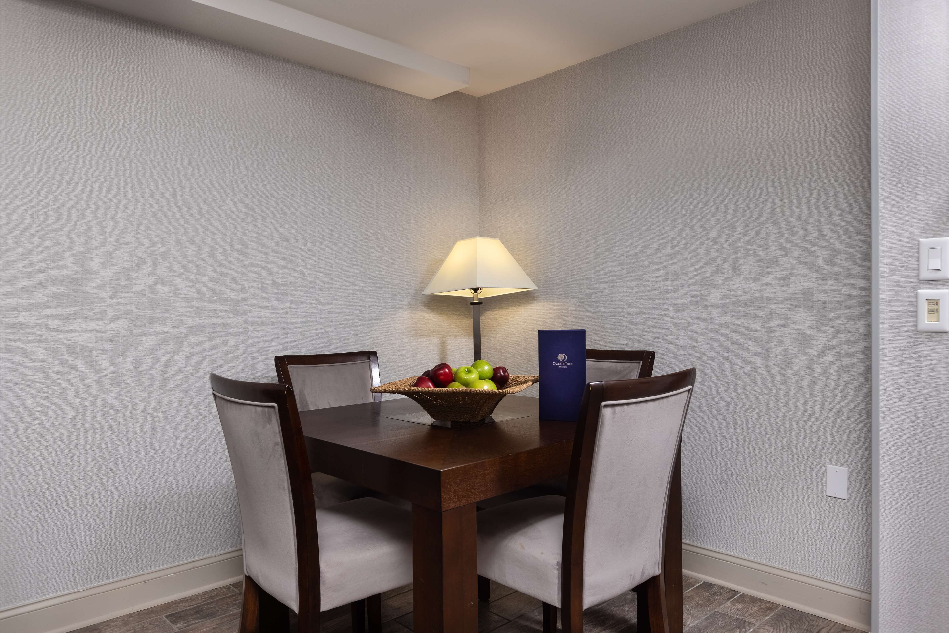 A dining table with four chairs, a basket of fruit, a lamp, and a blue folder on the table.