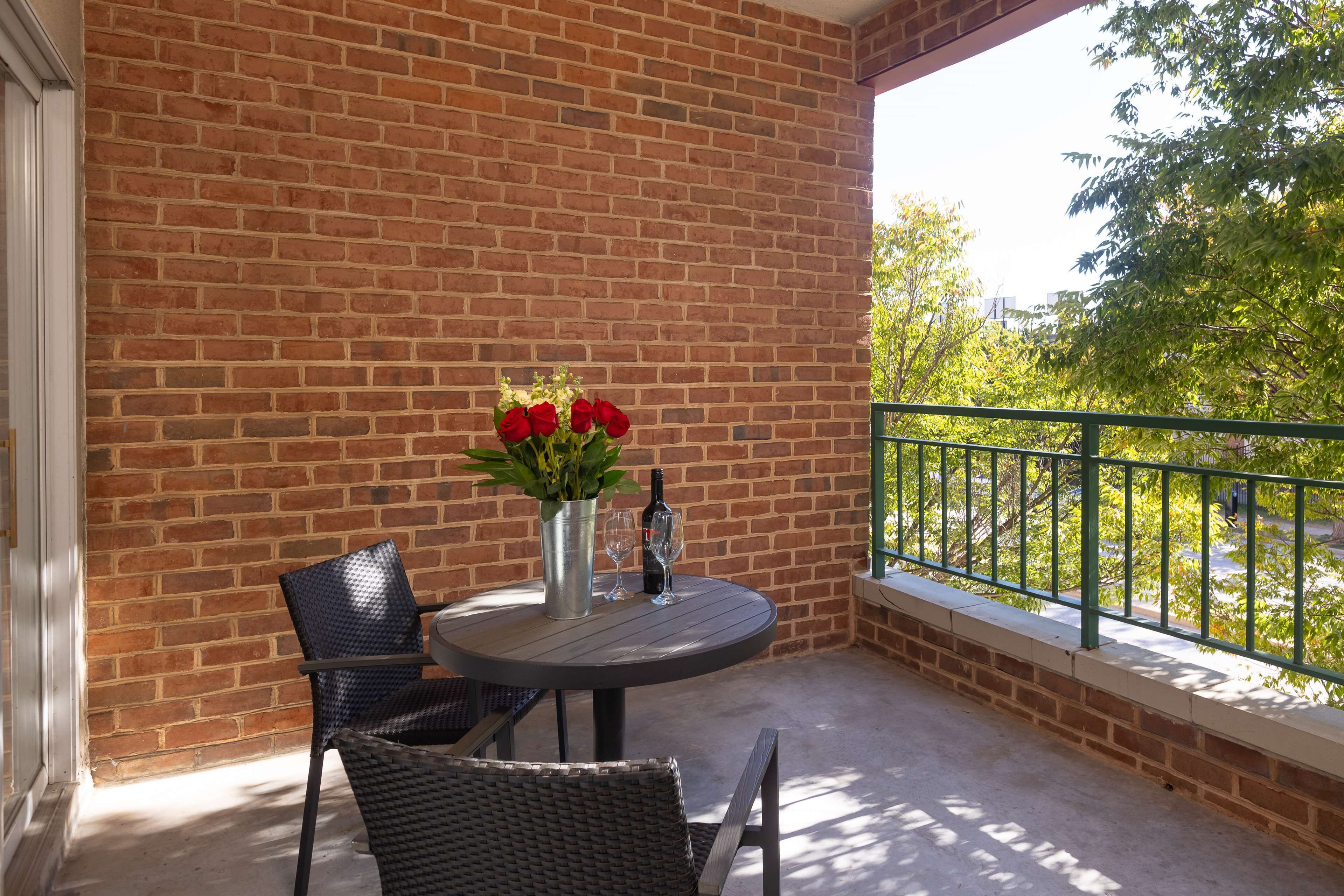 A brick wall, a round table with two chairs, a vase with red flowers, and a bottle.