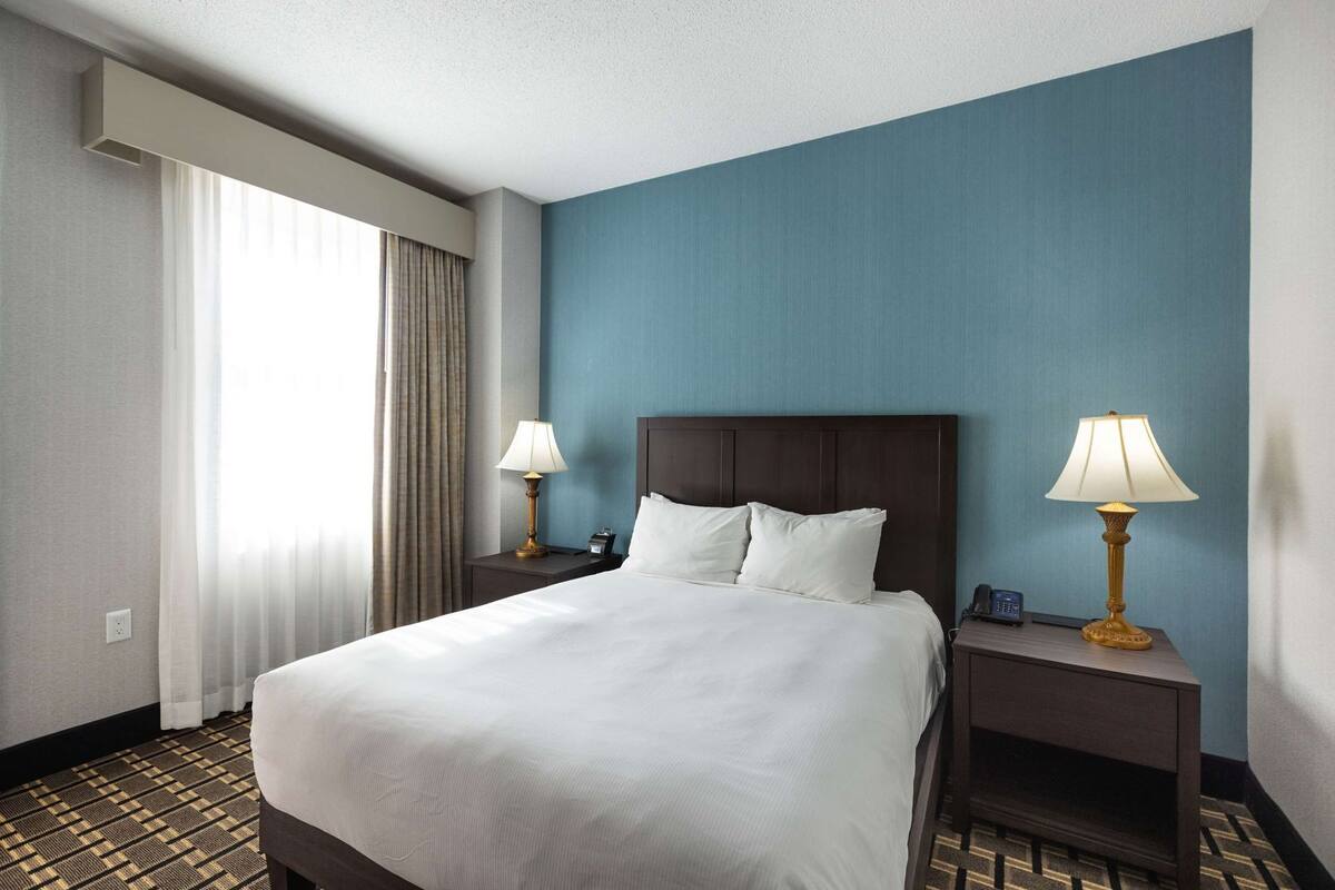 A hotel room with a large bed, two bedside tables with lamps, a window with curtains, and a patterned carpet.
