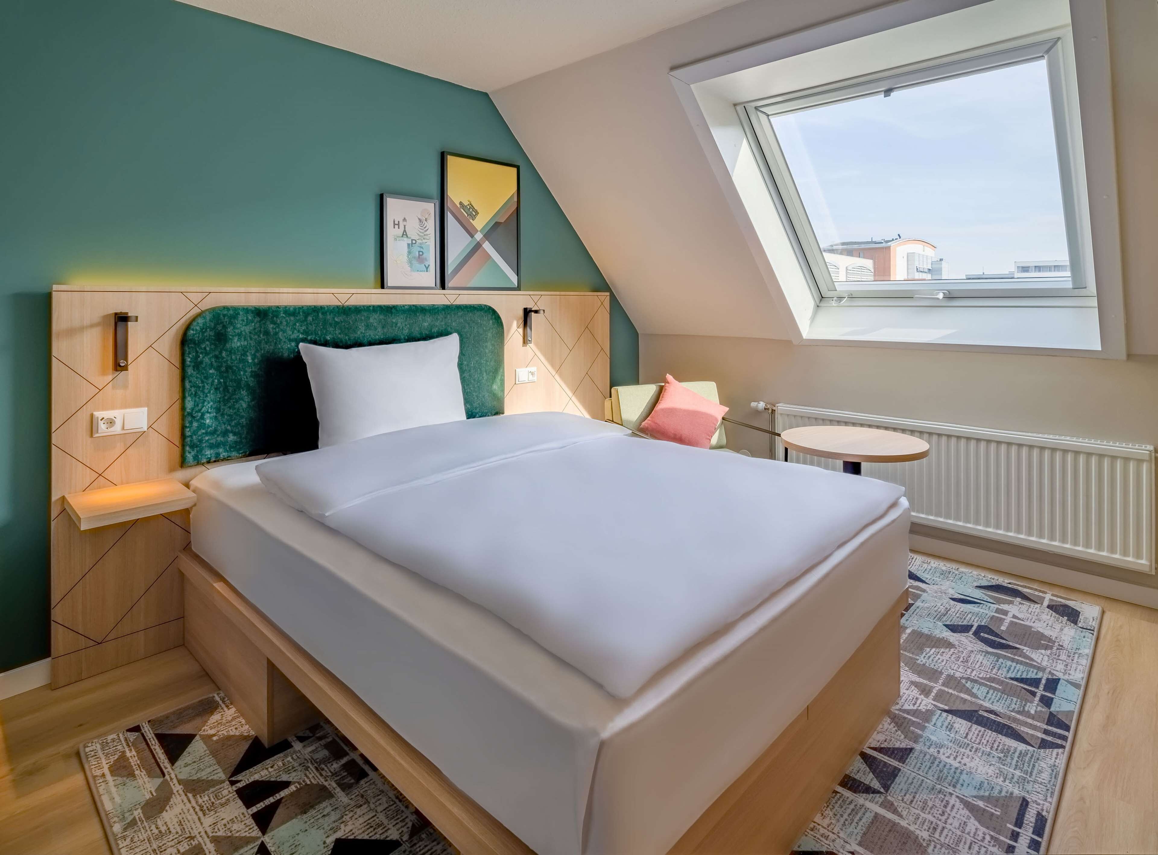 Una camera da letto moderna con un letto, un tavolino rotondo, una finestra e vista sugli edifici.