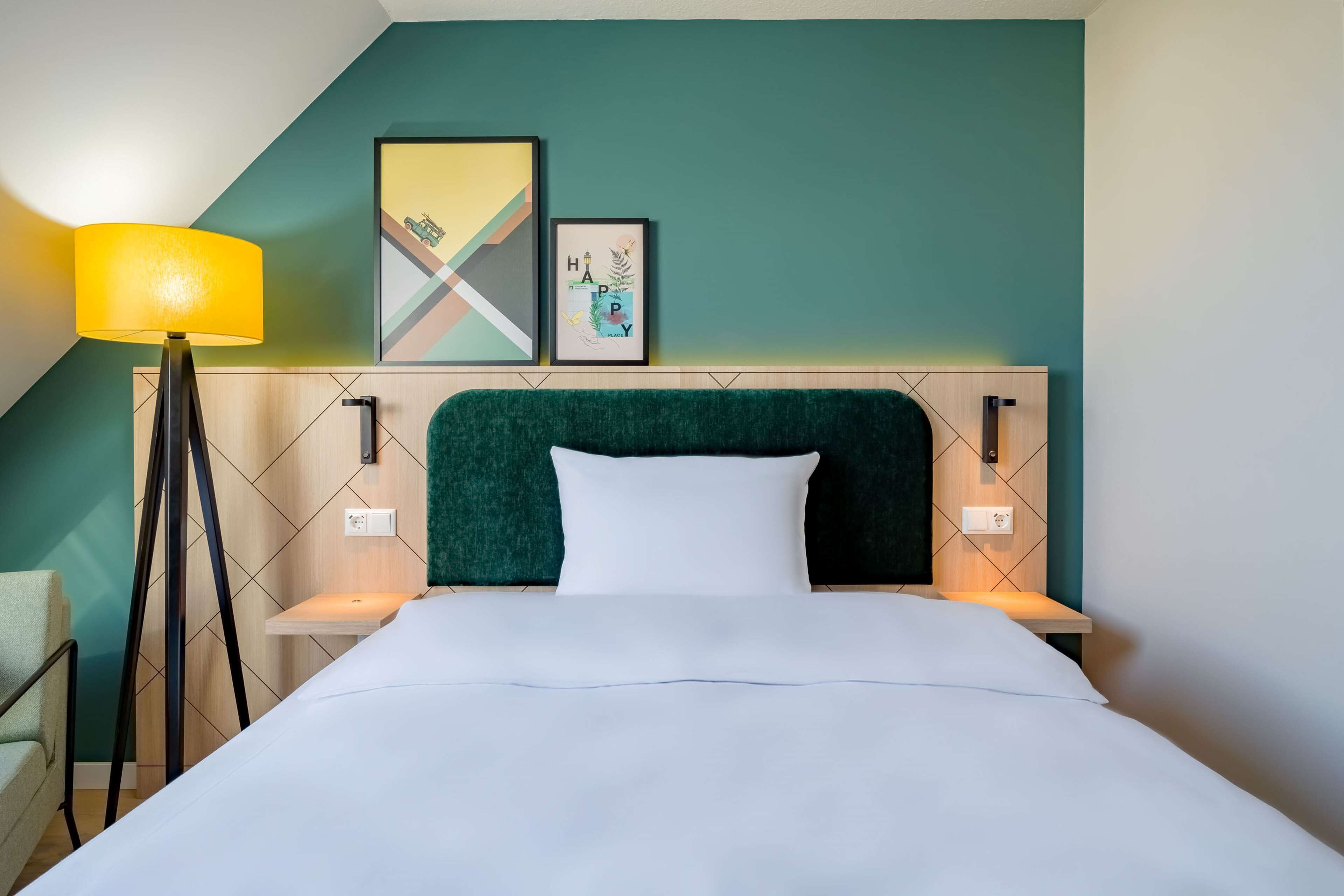 Una cama bien hecha con un cabecero verde, una lámpara de noche y dos cuadros en la pared.