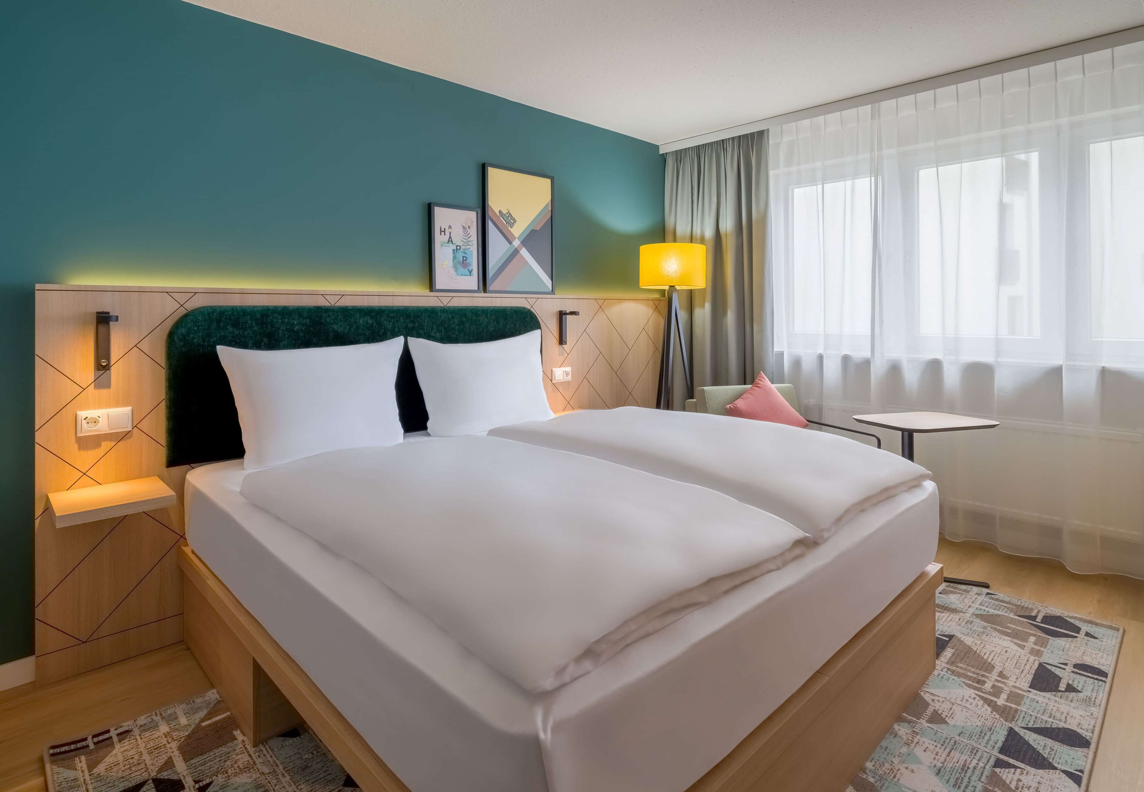 A double bed with white bedding, a headboard with a green upholstered section, two wall-mounted nightstands, a small table with a pink chair, a floor lamp, and a window with sheer curtains.