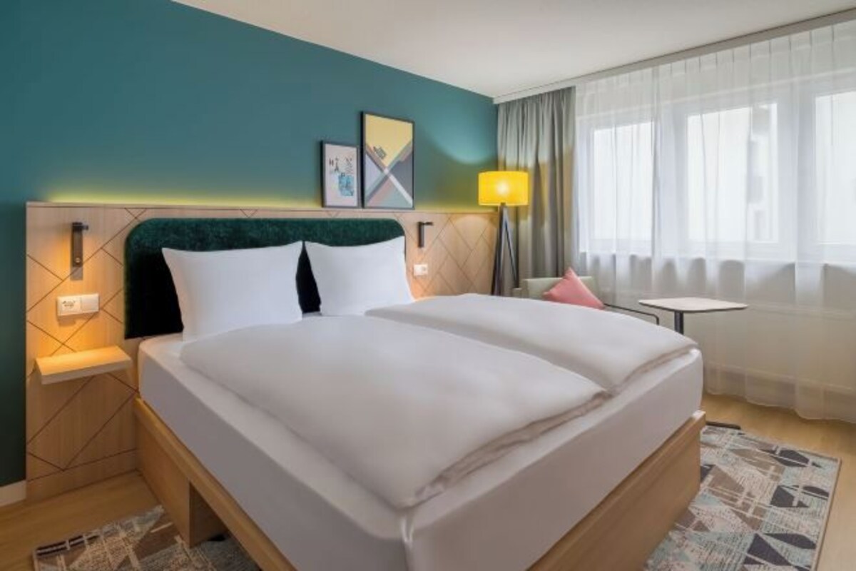 A hotel room with a bed, bedside tables, a lamp, a small table, and a window with curtains.