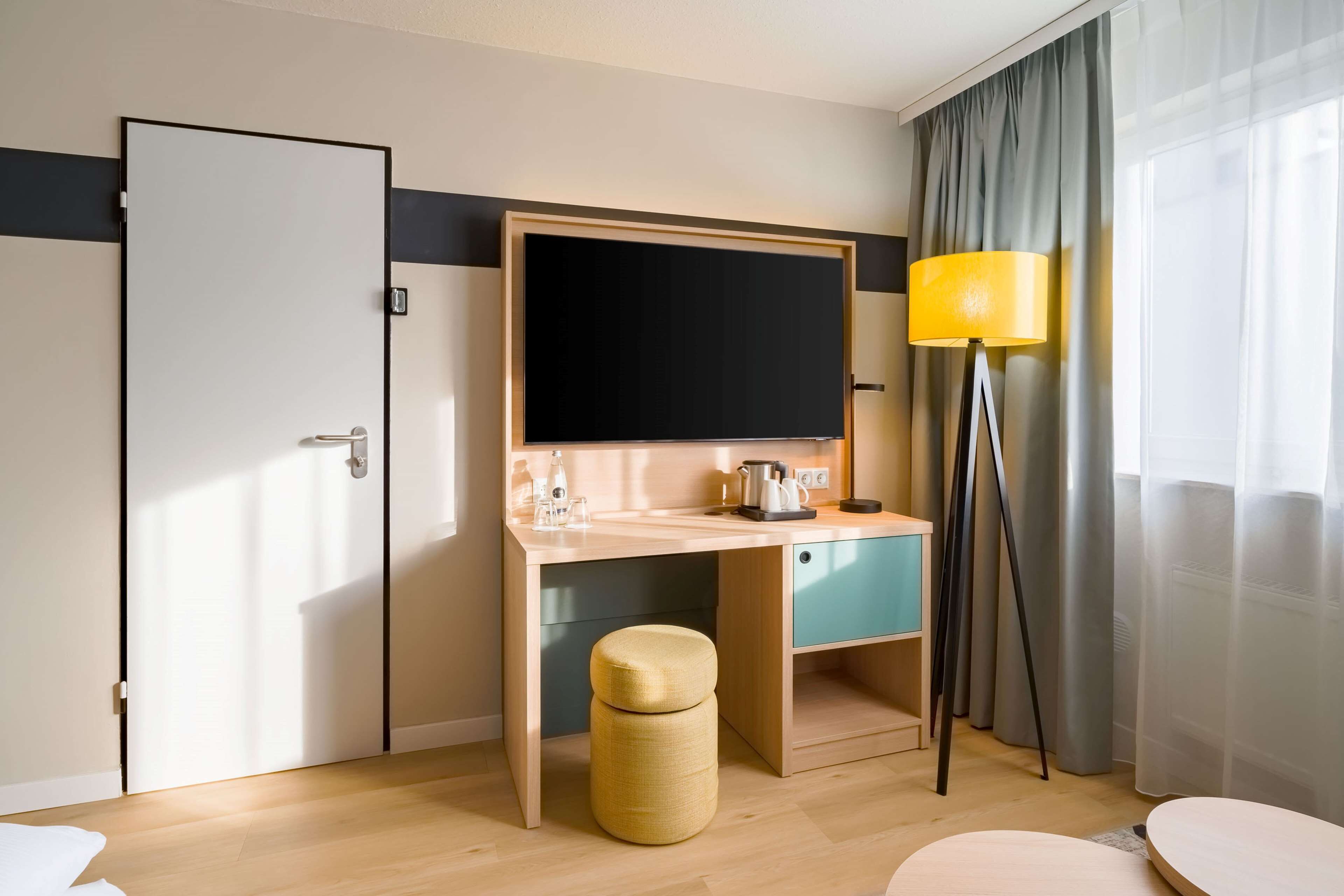 A hotel room with a wooden desk, a flat-screen TV, a lamp, and a small stool.