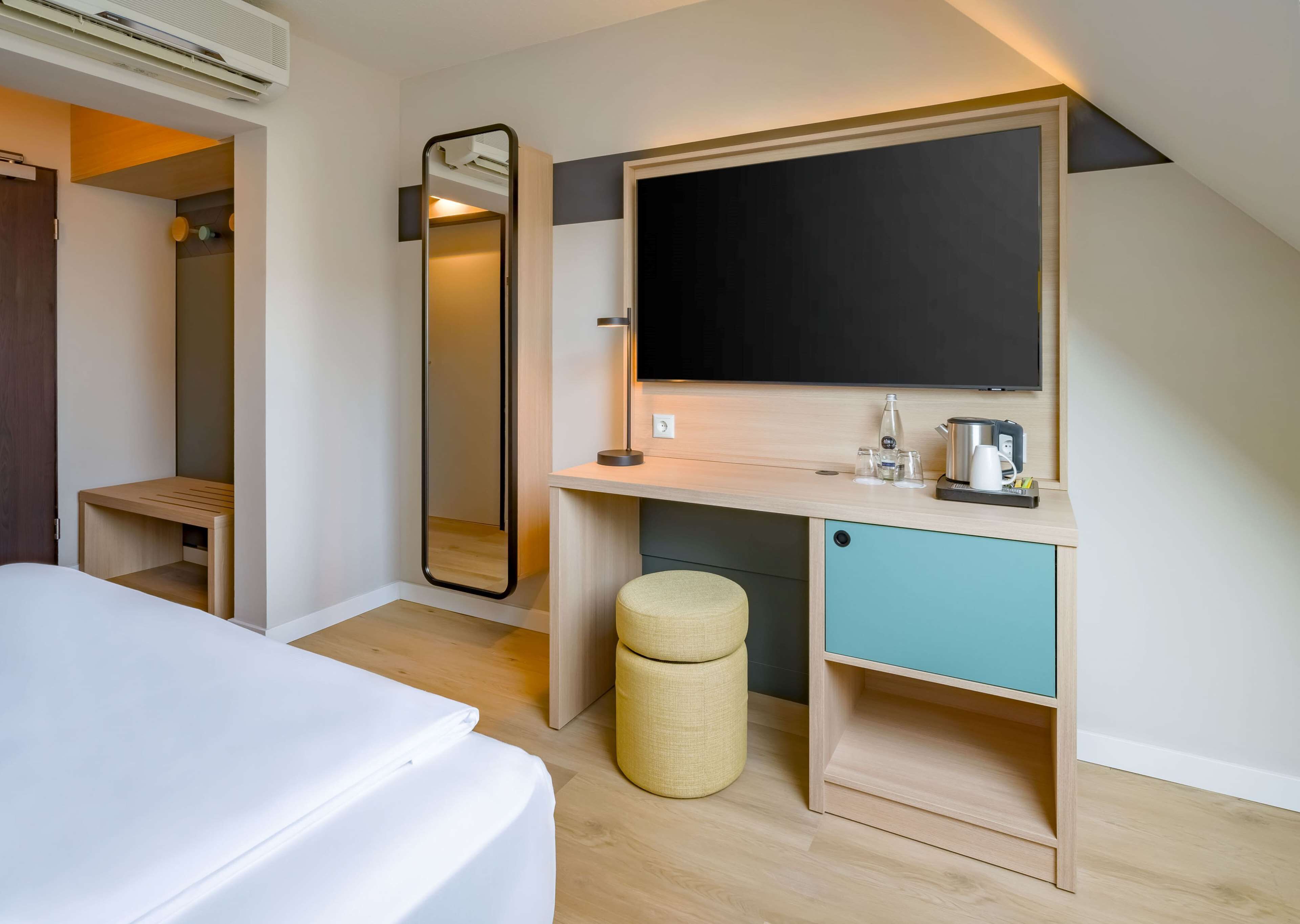 Een moderne hotelkamer met een bed, een houten bureau met een spiegel, een wastafel en een klein krukje.