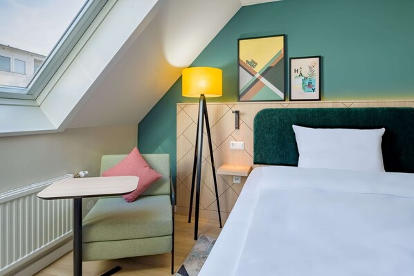 Ein Hotelzimmer mit Dachfenster, einem Bett, einem Schreibtisch, einem Stuhl, einer Stehlampe und Bildern an der Wand.