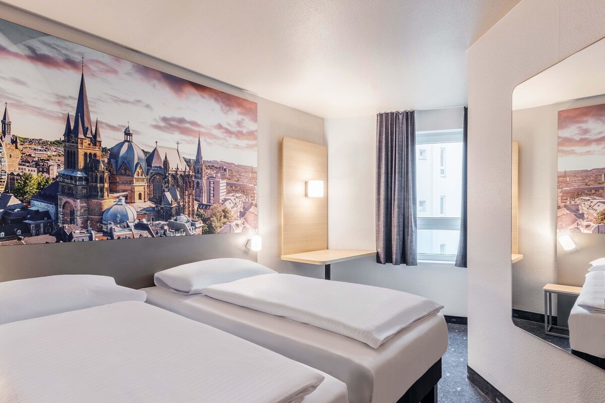 A hotel room with two beds, a cityscape mural, a window with curtains, and a small table.