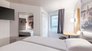 A hotel room with a bed, a desk, a chair, a mirror, and a window with curtains. - B&B Hotel Aachen-Hbf (Aachen)