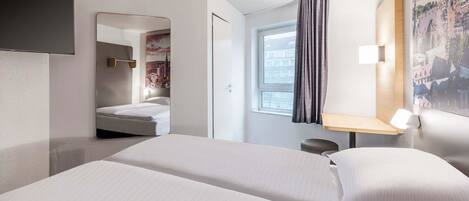 A hotel room with a bed, a desk, a chair, a mirror, and a window with curtains.