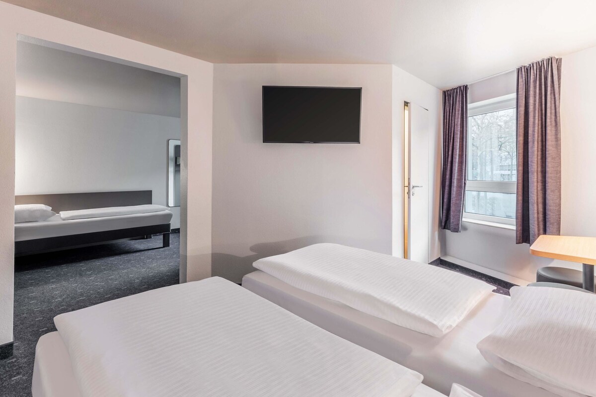 A hotel room with two single beds, a flat-screen TV mounted on the wall, and a window with curtains.