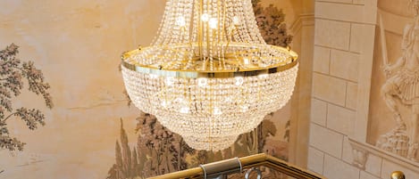 A grand staircase with a crystal chandelier, ornate wall murals, and a classic architectural design.