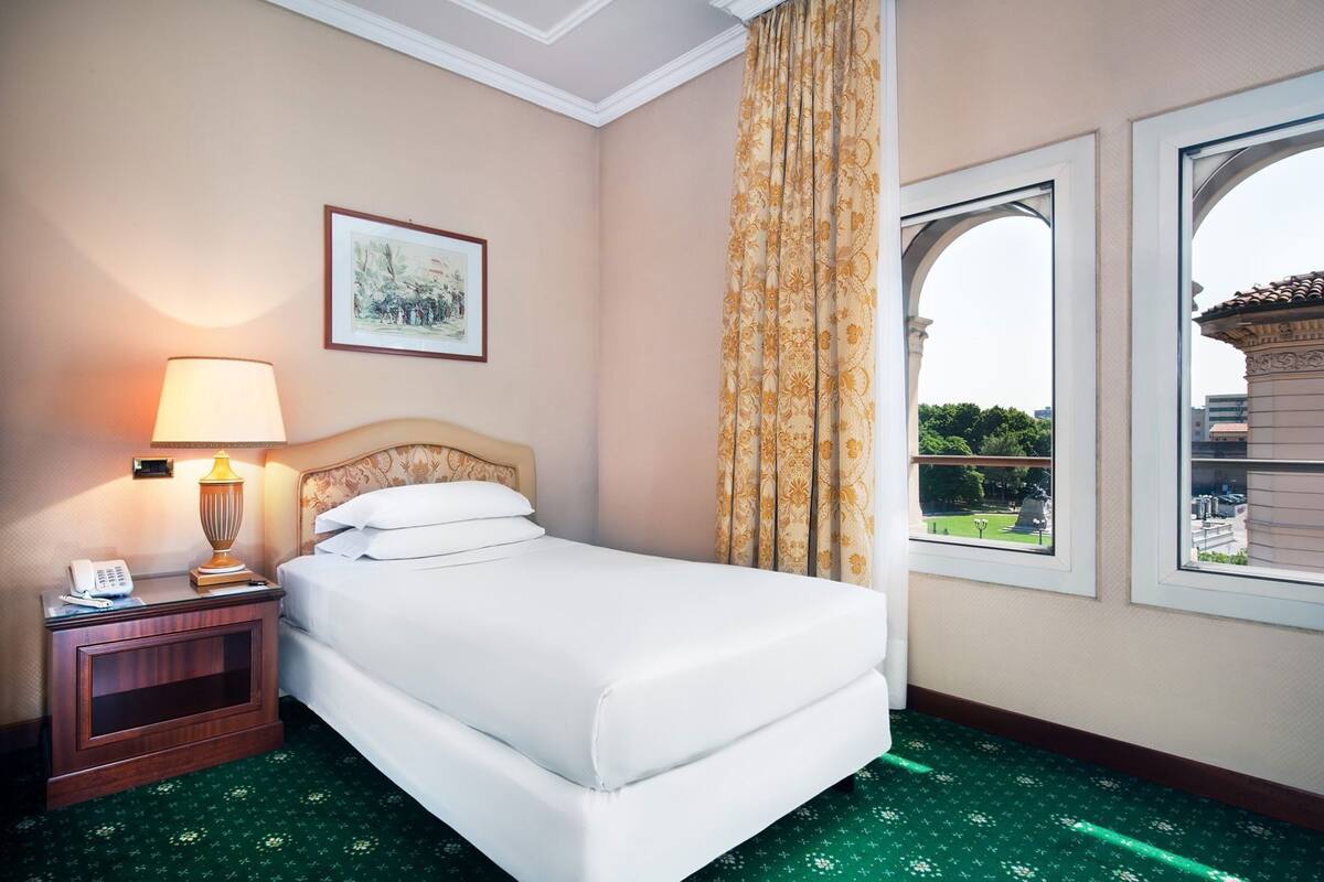 Une chambre d’hôtel avec un lit, une table de chevet avec une lampe, un tableau encadré au mur et une fenêtre donnant sur un paysage verdoyant et un bâtiment.