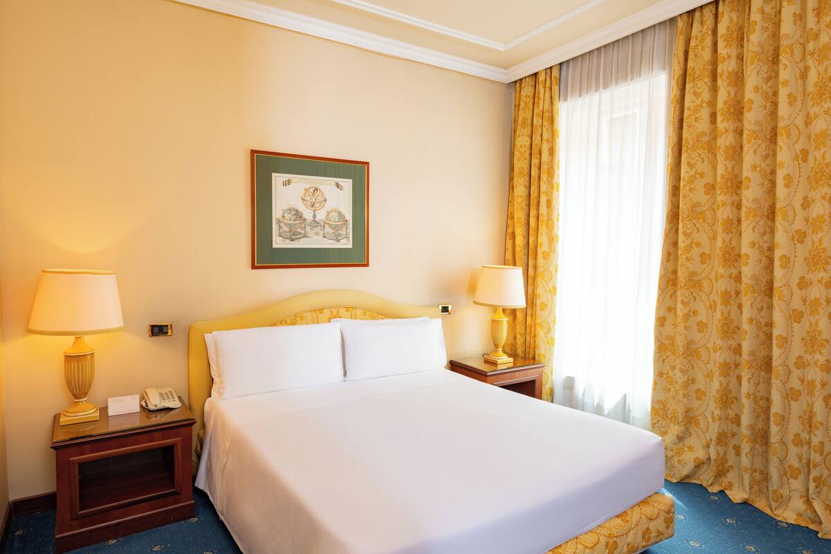 Une chambre d’hôtel avec un lit, deux tables de chevet avec des lampes, un tableau encadré au mur et une fenêtre avec des rideaux.