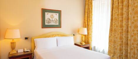 A hotel room with a bed, two bedside tables with lamps, a framed picture on the wall, and a window with curtains.
