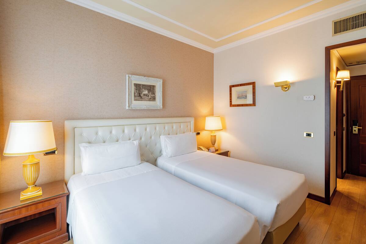 Une chambre d’hôtel avec deux lits, une table de chevet avec une lampe, un tableau encadré au mur et une porte donnant sur une autre pièce.
