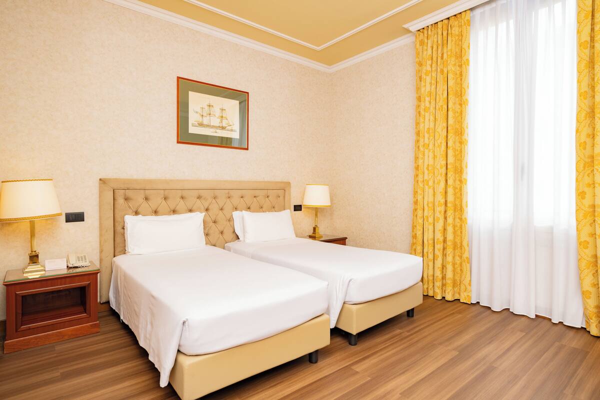 Une chambre d’hôtel avec deux lits, un tableau encadré au mur et une fenêtre avec des rideaux jaunes.
