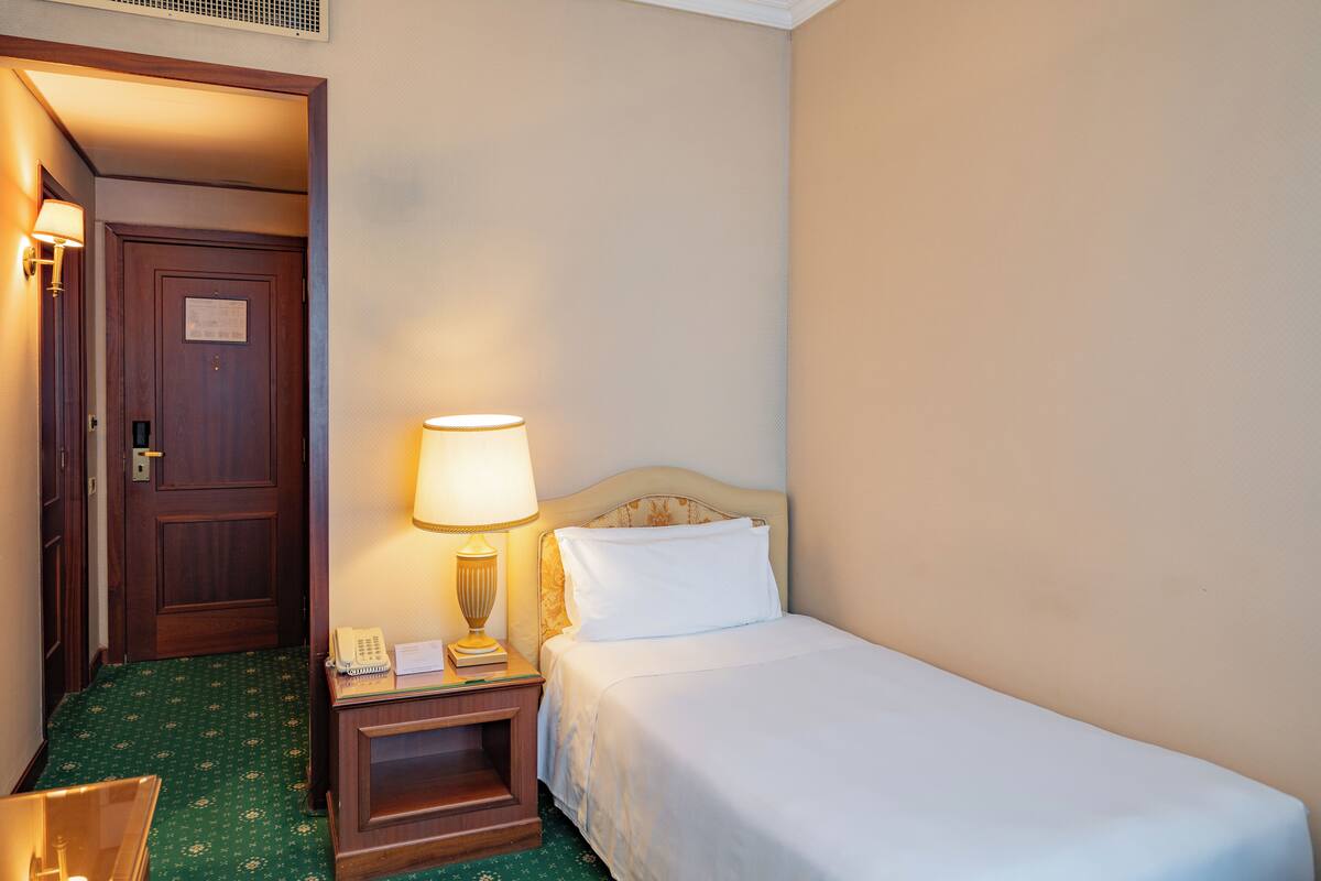 Une chambre d’hôtel avec un lit simple, une table de chevet avec une lampe, un téléphone et une porte.