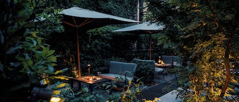 Un patio al aire libre con mobiliario y sombrillas, rodeado de abundante follaje.