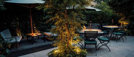 Ein Terrassenbereich im Freien mit Tisch und Stühlen, umgeben von Grünpflanzen und Bäumen.