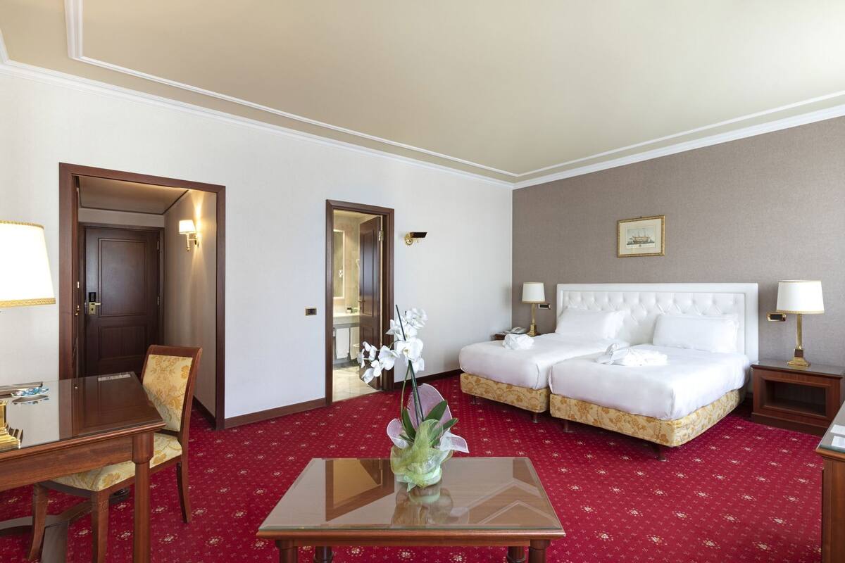 Une chambre d’hôtel avec un lit double, un bureau avec une chaise, une petite table ornée d’un vase de fleurs et un sol recouvert d’une moquette rouge.
