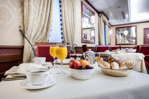 Une table mise pour le petit-déjeuner, avec une nappe pour deux personnes, comprenant un saladier de fruits, des viennoiseries et deux verres de jus d’orange.