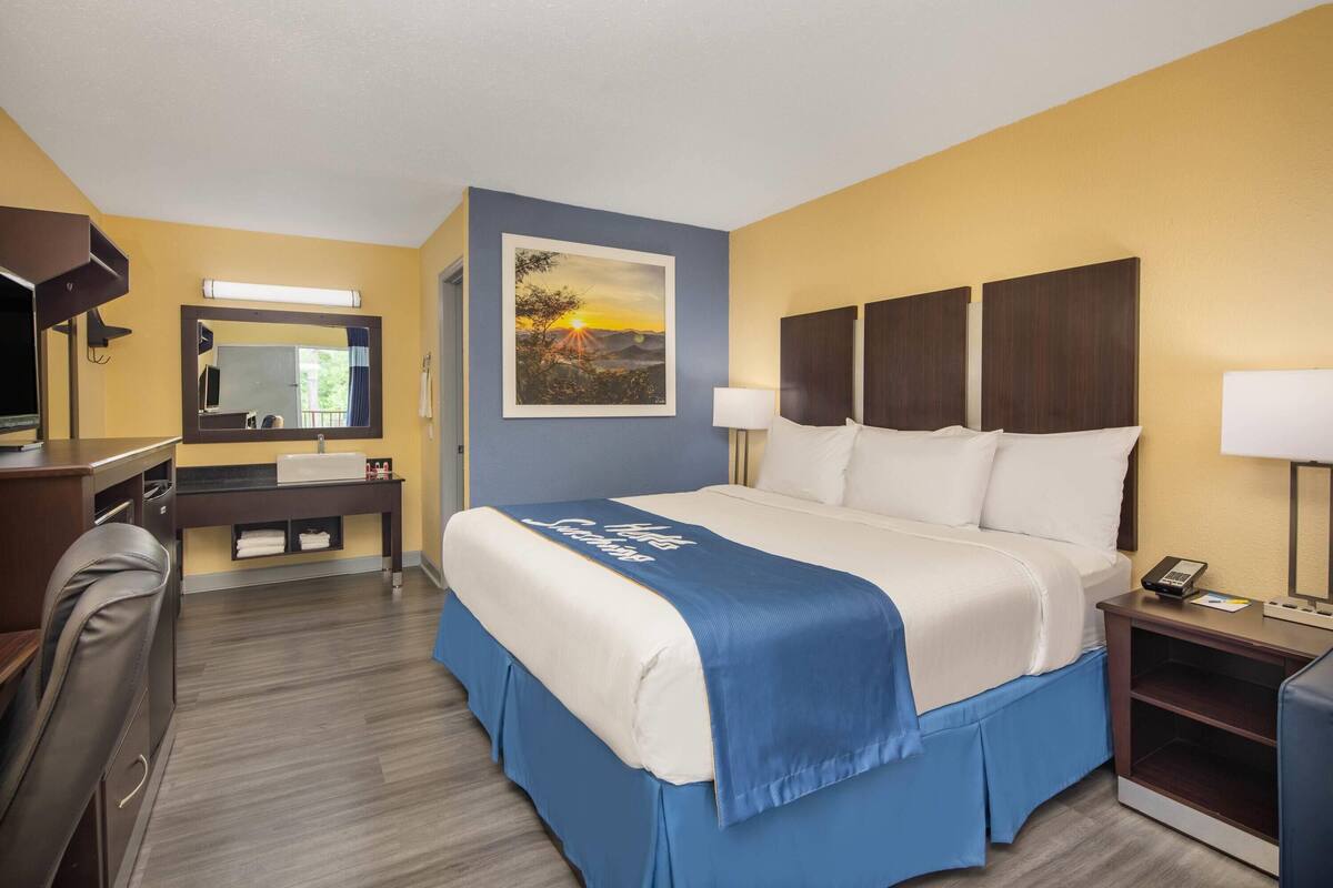 Habitación de hotel con una cama, un escritorio, una silla, un espejo, un cuadro y una mesita de noche con una lámpara.
