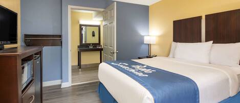 A hotel room with a bed, a desk with a lamp, a TV, and a bathroom visible through an open door.