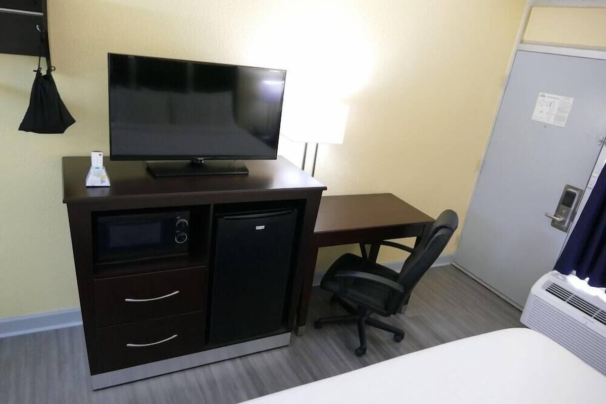 Habitación de hotel con cama, escritorio con televisor, refrigerador, microondas, silla y puerta con cerradura.