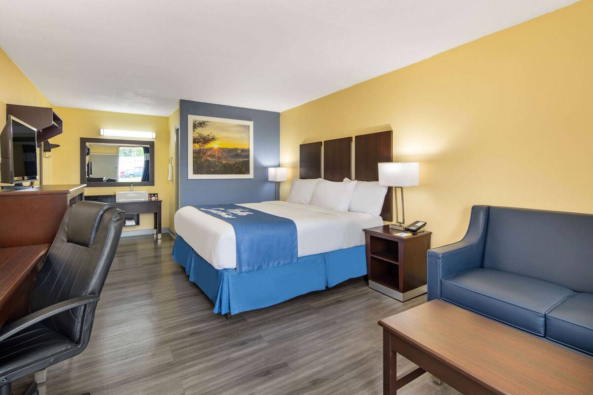 Una habitación de hotel con cama, escritorio, silla, sofá y mesita de noche con lámpara.