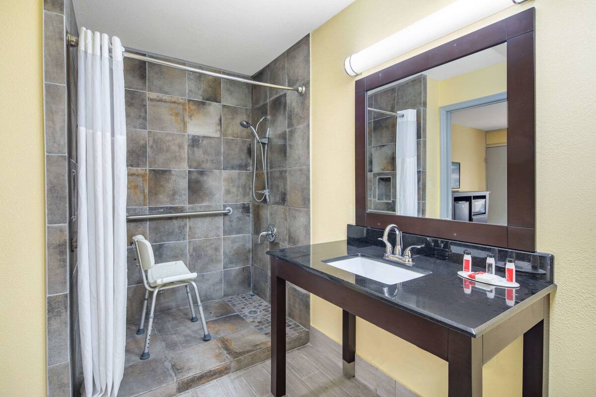 Un baño con ducha, una silla accesible para sillas de ruedas, un lavamanos con espejo y un perchero para toallas.