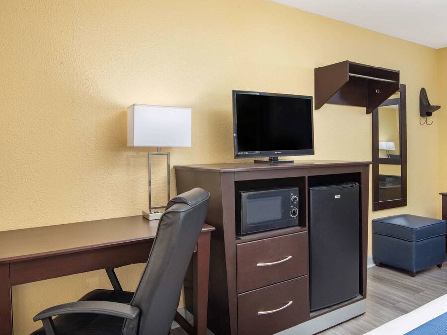 Quarto de hotel com escrivaninha, televisão, micro-ondas, frigorífico, cadeira, mesa pequena e espelho.