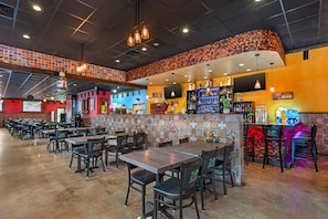 Interior de un restaurante con varias mesas y sillas, una barra con estantes llenos de botellas y una pared de azulejos de colores.