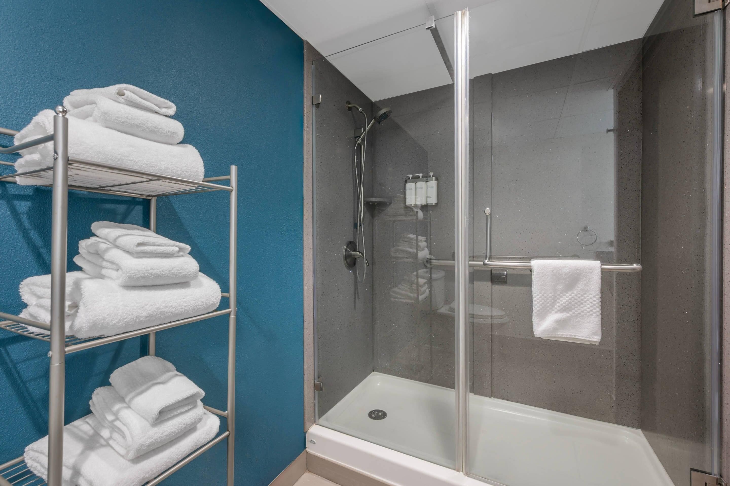 A glass-enclosed shower stall with a white bathtub and a metal towel rack with folded towels.