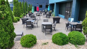 Une terrasse extérieure avec des tables et des chaises, entourée d’arbustes soigneusement taillés et, au fond, d’un bâtiment bleu.