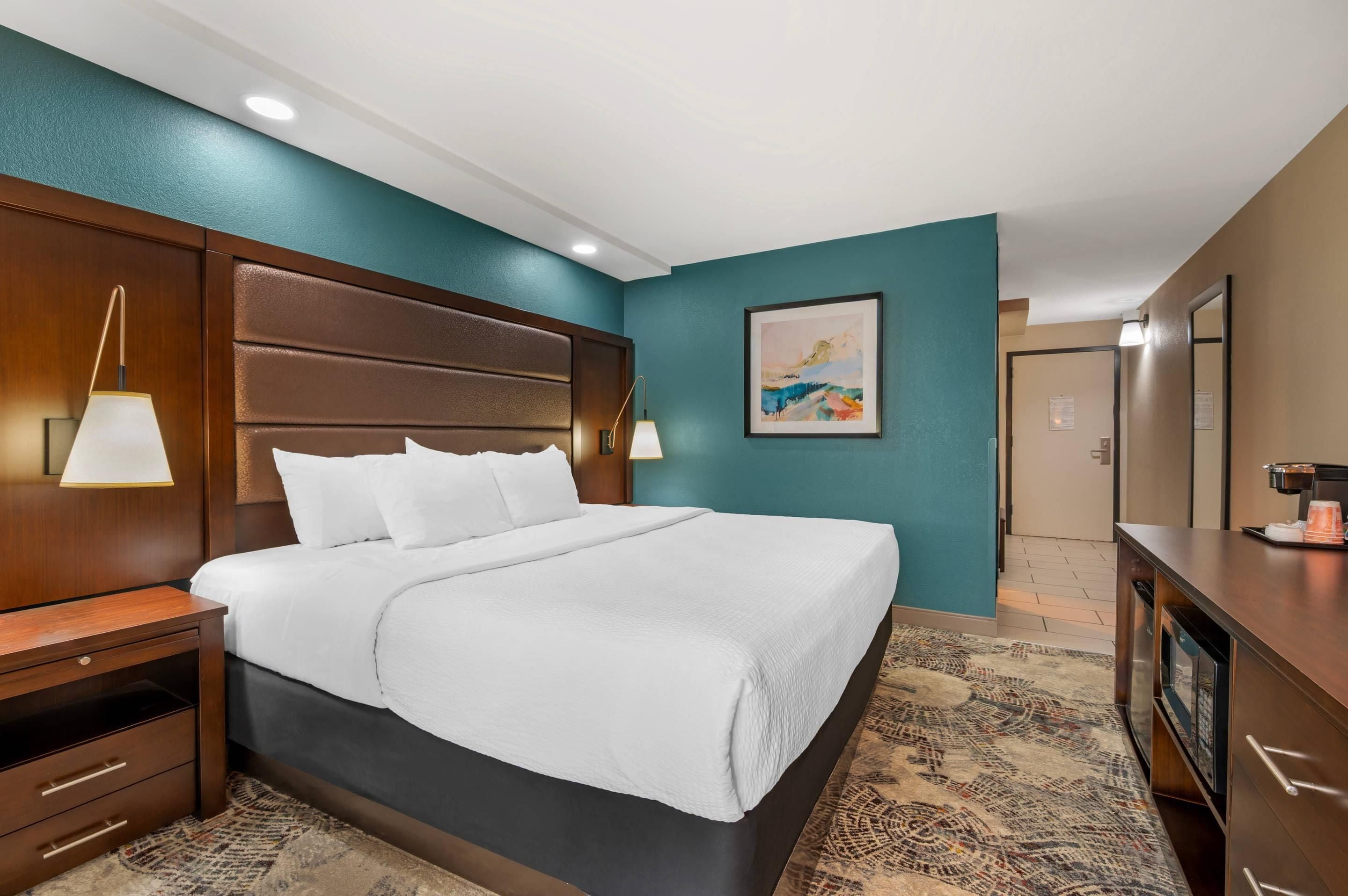 Habitación de hotel con una cama grande, cabecero de madera, dos lámparas de noche, una obra de arte enmarcada en la pared y un escritorio con cafetera.