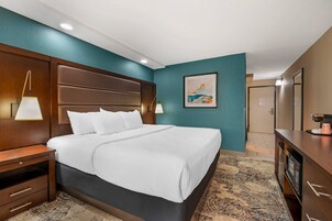 Habitación de hotel con una cama grande, cabecero de madera, dos lámparas de noche, una obra de arte enmarcada en la pared y un escritorio con cafetera.