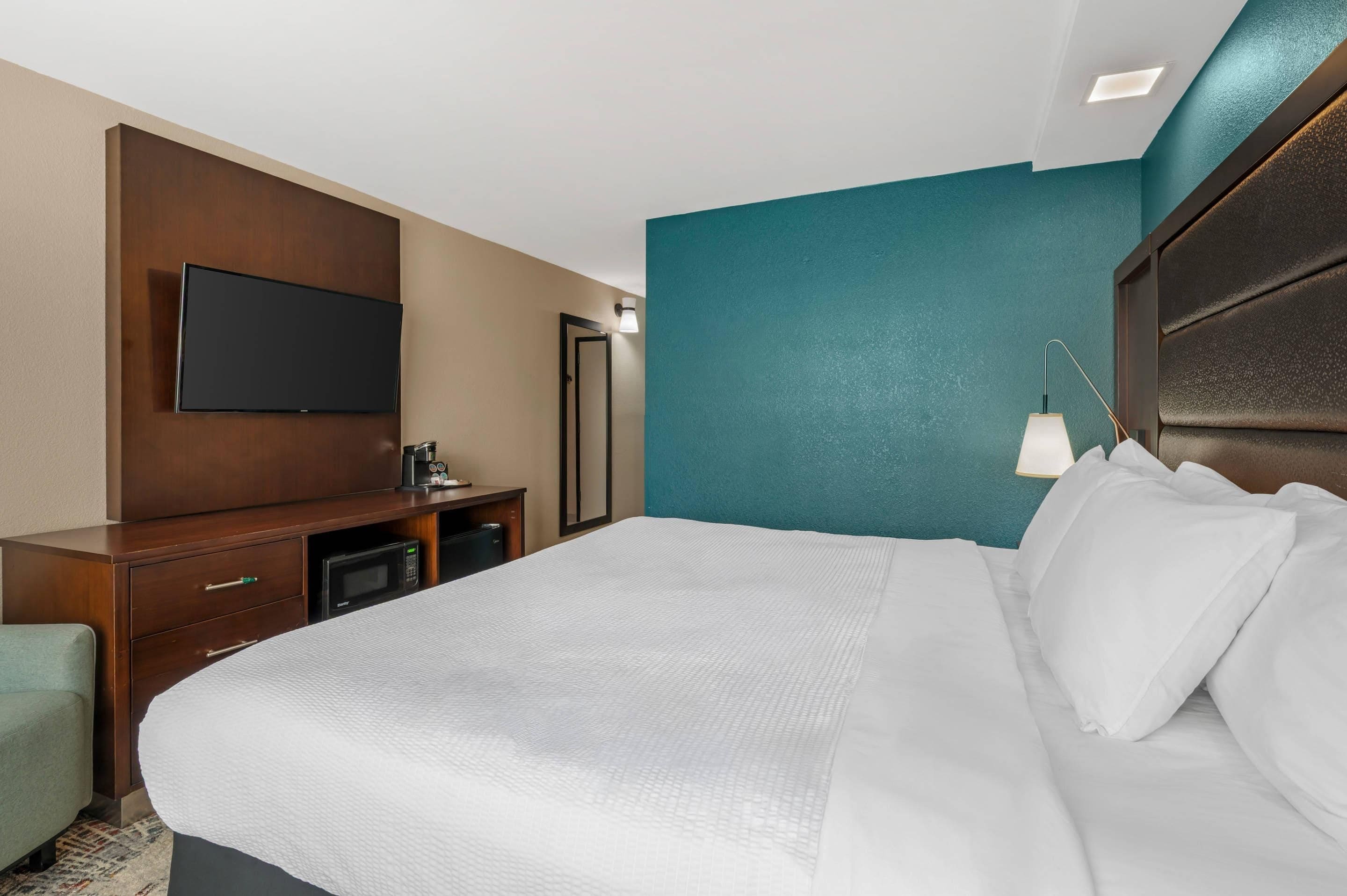 Una camera d'albergo con un letto, una TV a schermo piatto fissata al muro, una scrivania con una macchina del caffè e una lampada.