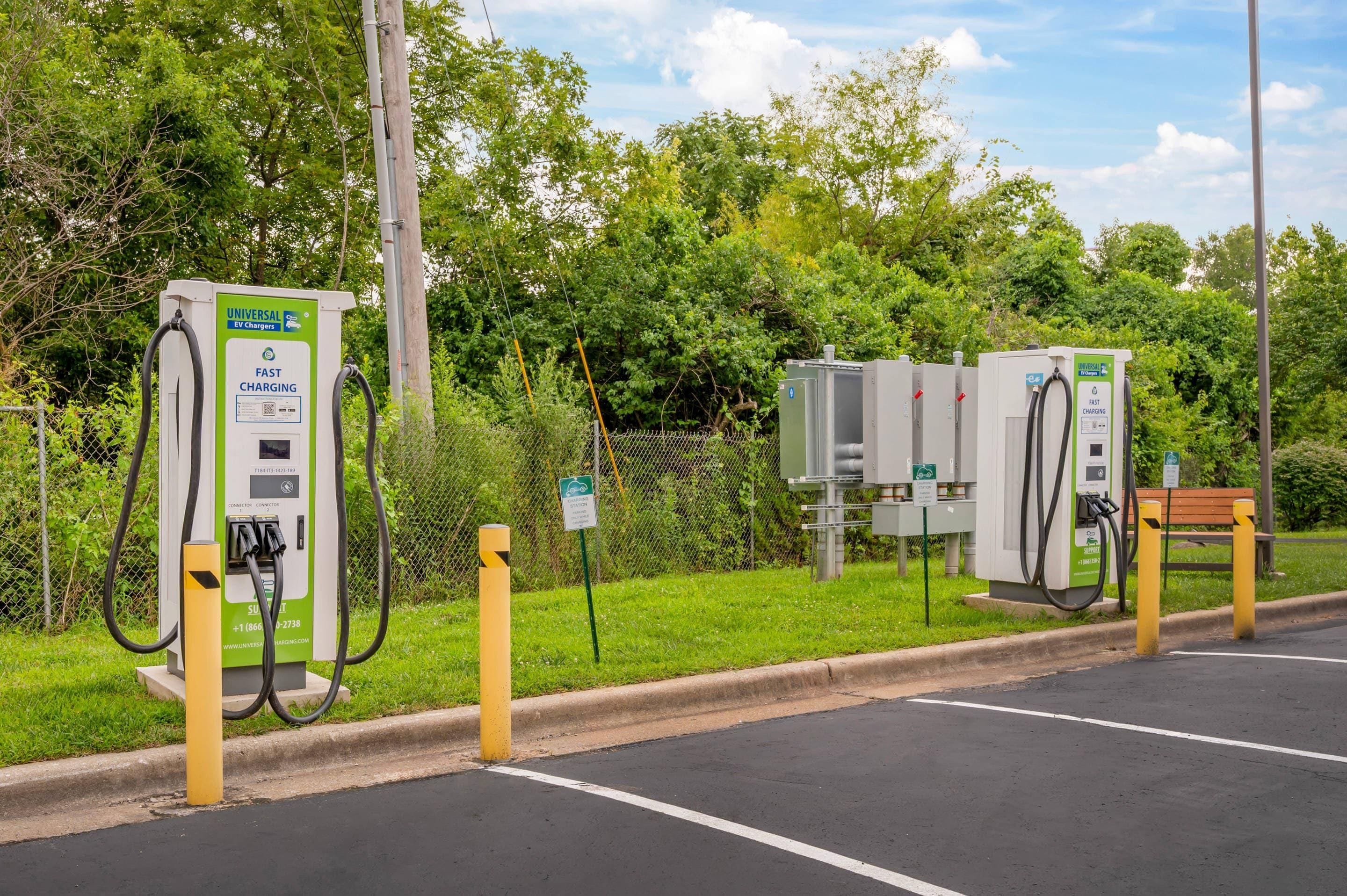 Un estacionamiento con varias estaciones de carga para vehículos eléctricos, un banco y una cerca de malla metálica.