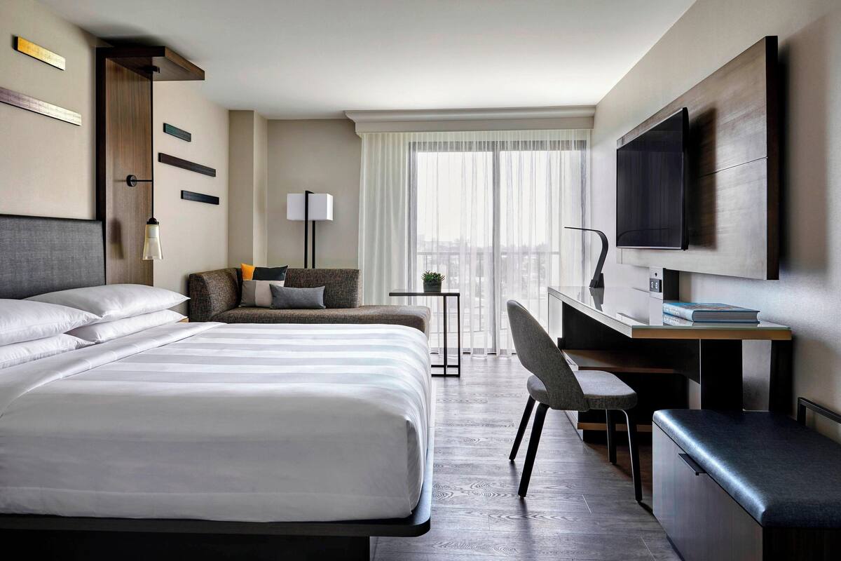 A modern hotel room with a large bed, a desk, a chair, and a TV.